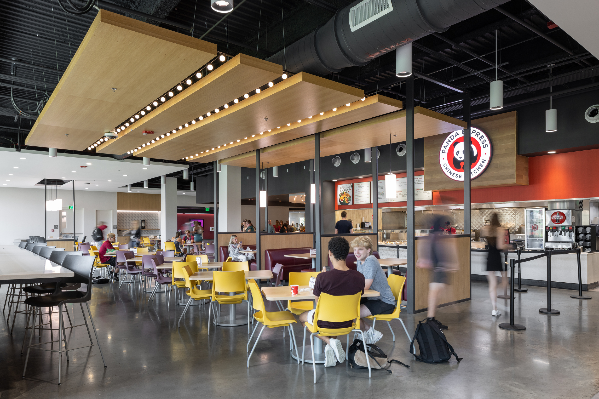 View of the inside of the Florida State University Student Union food court. The interior has an industrial, modern design with wooden accents and stylish edison bulbs. College kids are sitting at dining tables with bright yellow chairs eating their meals. In the background is a Panda Express restaurant.